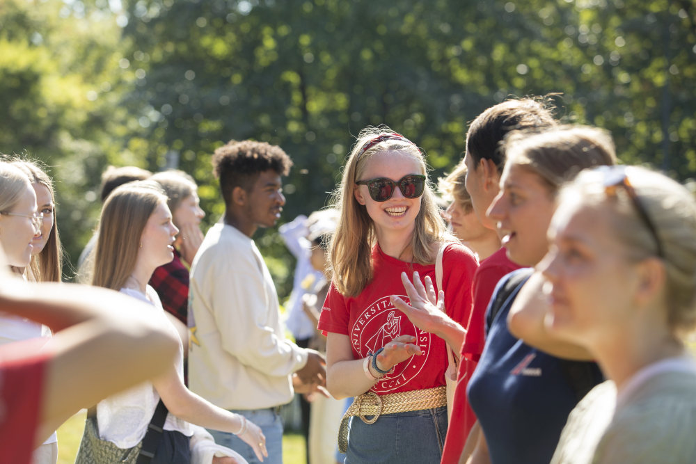 Syv studenter i r?d fadder-t-skjorte st?r med ryggen mot kamera, noen holder opp armen. Solfylt dag p? Blindern i bakgrunnen.